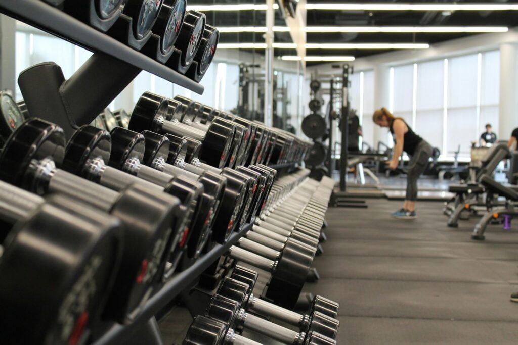 A woman lifts weights behind a dumbbell rack in a commercial gym.